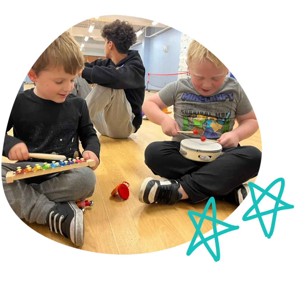 Two young children playing musical instruments together on a wooden floor during a music activity, smiling and focused.